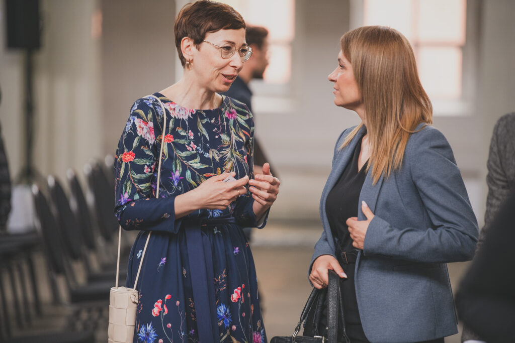 Zdjęcie przedstawia dwie rozmawiające kobiety. W tle widoczne są rzędy krzeseł.