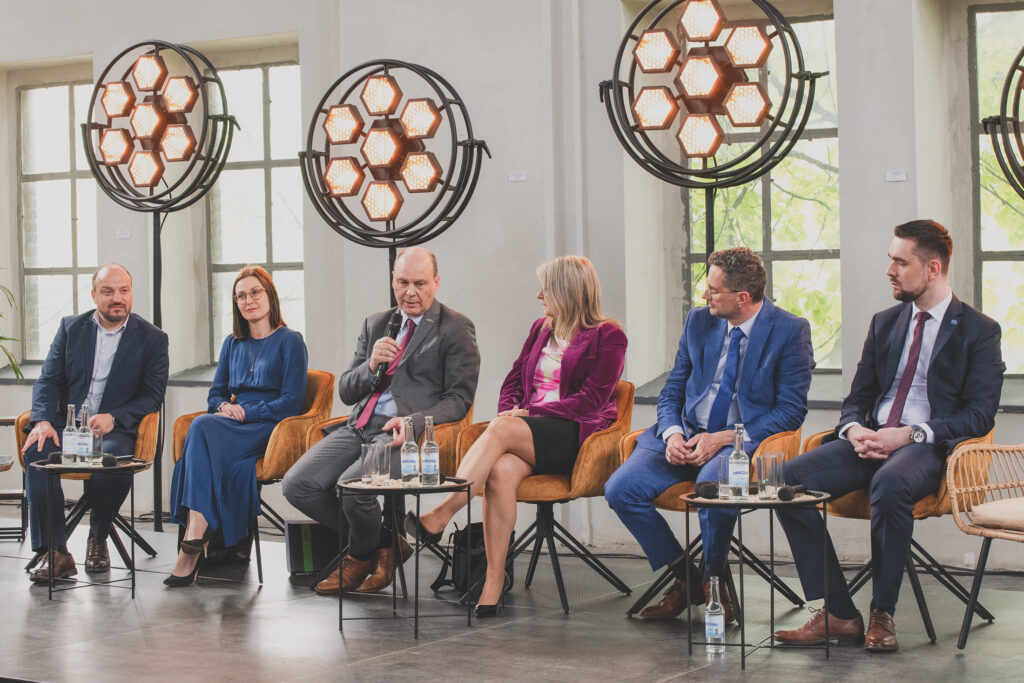 Zdjęcie przedstawia dwie kobiety oraz czterech mężczyzn w biznesowych strojach, siedzących w rzędzie na miękkich, brązowych fotelach. Za nimi znajduje się rząd lamp oraz okien. Jeden z mężczyzn trzyma w dłoni mikrofon.