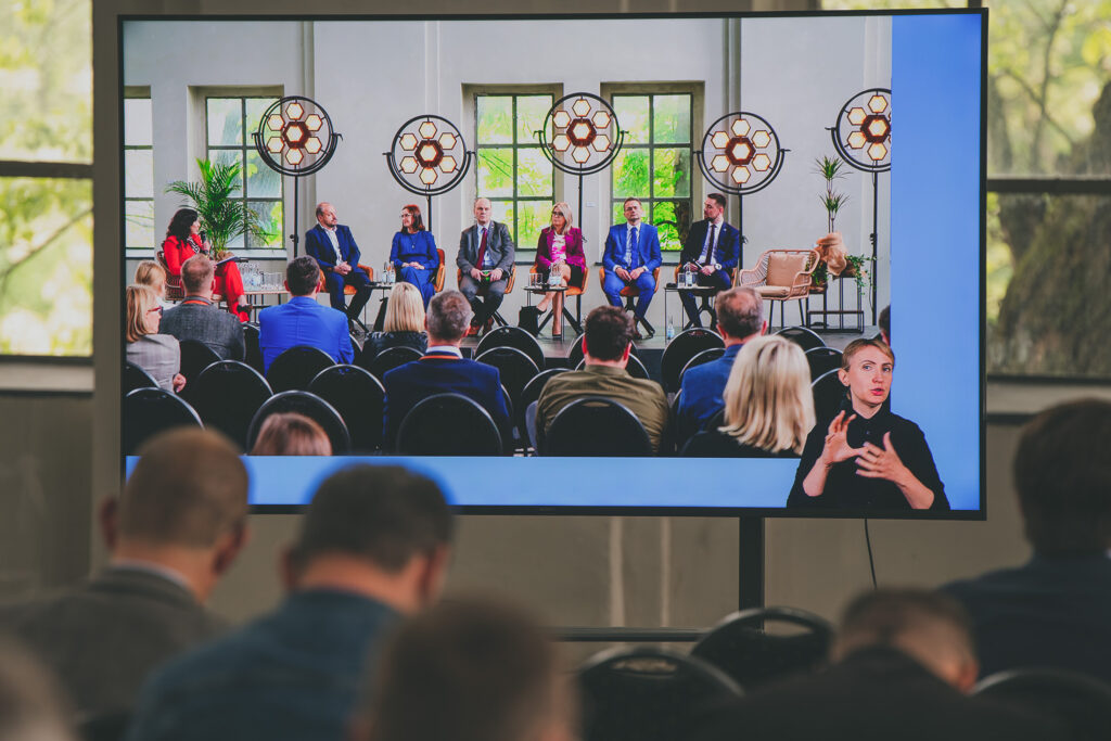Zdjęcie przedstawia fragment wnętrza sali w trakcie konferencji. Na pierwszym planie widoczne są głowy siedzących osób, a na tle okien znajduje się ogromny ekran, na którym transmitowana jest konferencja. Na ekranie widać prelegentów oraz moderatorkę dyskusji, a także kobietę tłumaczącą ich słowa na język migowy.