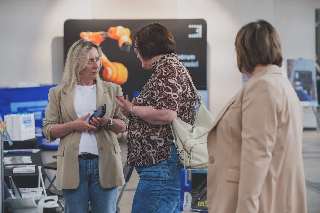 Zdjęcie przedstawia trzy kobiety. W tle widoczne są ścianki reklamowe instytucji oraz torby z motywem przewodnim Funduszy Europejskich.