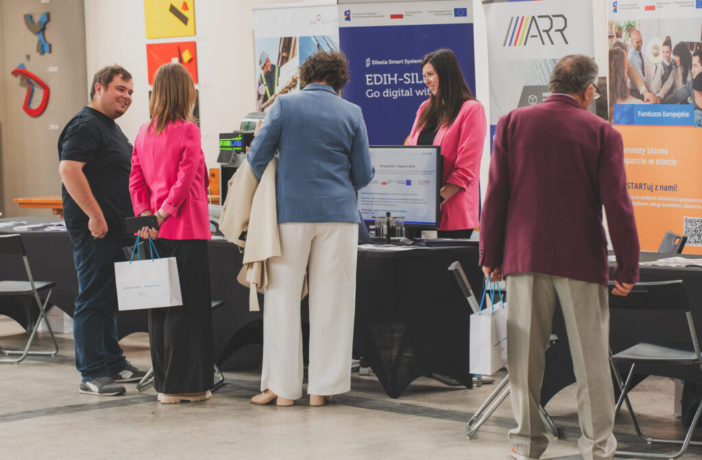 Za stolikiem przykrytym czarną tkaniną stoi kobieta w różowej marynarce. Roll-up za nią zawiera logo Agencji Rozwoju Regionalnego. Przed stolikami stoi grupa osób. Jeden z mężczyzn uśmiecha się do stojącej przy nim kobiety.