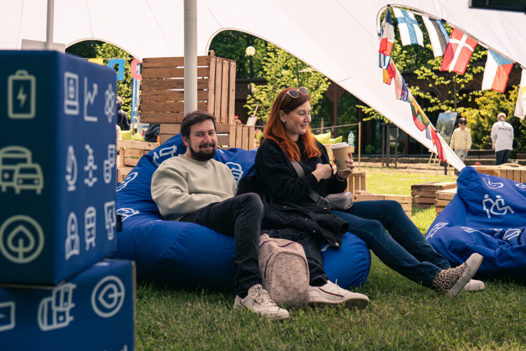 Na ogromnym pufie sako siedzą uśmiechnięci kobieta i mężczyzna. W tle widoczne są stalugi ze zdjęciem oraz chłopiec i animator.