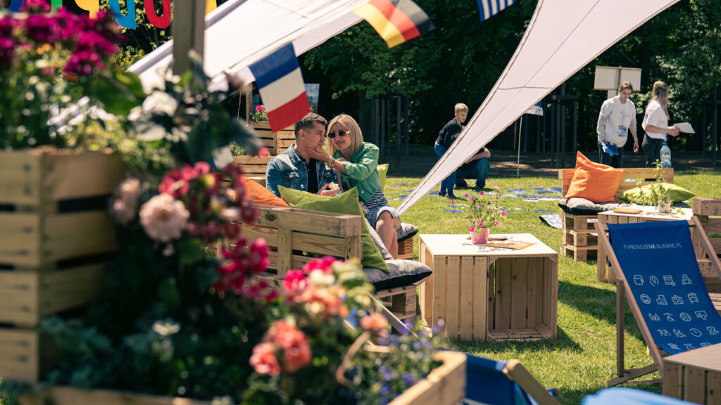 Na przykrytych poduszkami europaletach siedzi para. Kobieta głaszcze mężczyznę po policzku. Na pierwszym planie widoczne są skrzynki z kwiatami oraz leżak z napisem funduszeUE.slaskie.pl, a tle drzewa i inni uczestnicy pikniku.