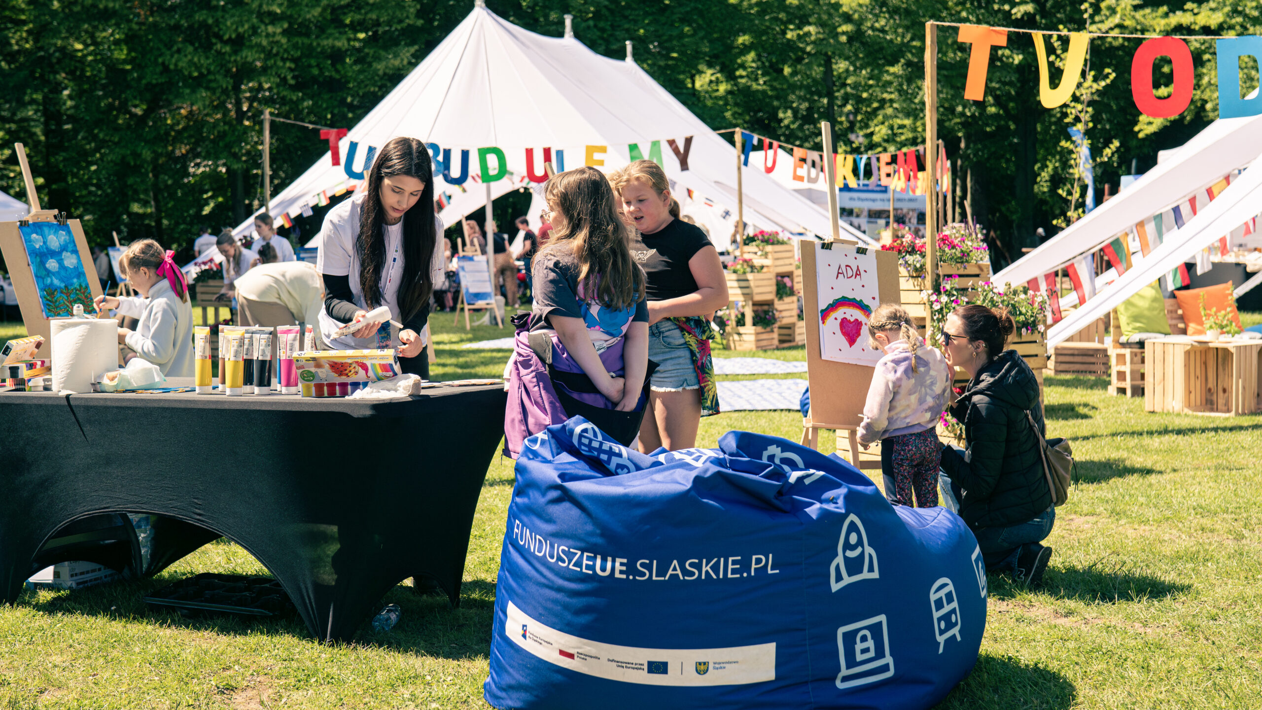 Zdjęcie przedstawia piknik. Na pierwszym planie widoczny jest puf sako z napisem funduszeUE.slaskie.pl. Za nim stoi stół zastawiony farbami, przy którym stoją animatorka oraz dwie dziewczynki. Na kolejnym planie stojąca przy sztalugach dziewczynka maluje niebo oraz rośliny. Przy innej sztaludze kobieta pomaga małej dziewczynce malować tęczę i serce, nad którym umieszczone jest imię Ada. W tle widoczne są drzewa, namioty i rozwieszone napisy: Tu budujemy, tu edukujemy.