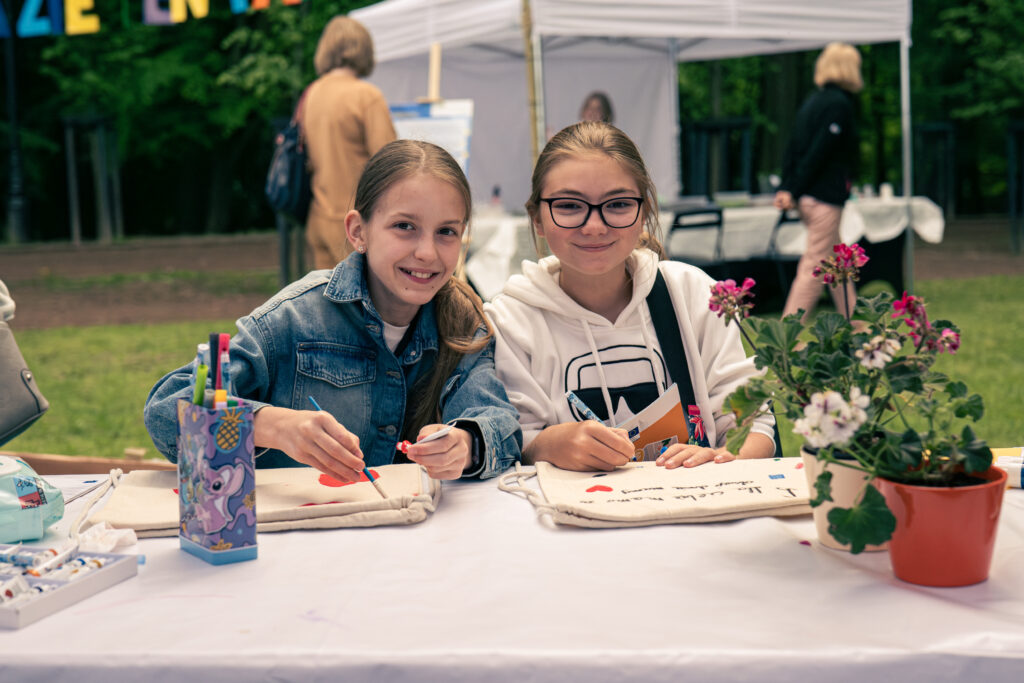 Przy stole siedzą uśmiechnięte dziewczynki ozdabiające ekologiczne plecaki. Obok stoją pelargonie w doniczkach. W tle widoczne są drzewa, namiot oraz inni uczestnicy pikniku.