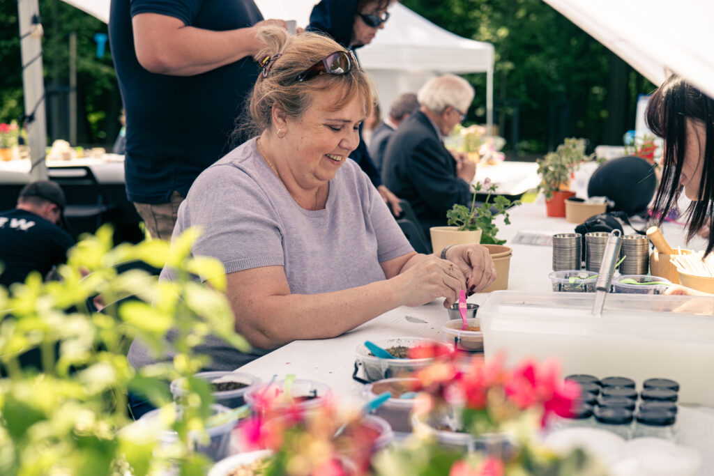 Uśmiechnięta kobieta tworzy coś przy stole. Na stołach stoją metalowe miseczki, różne składniki w małych wiaderkach, a także kwiaty i zioła w doniczkach.