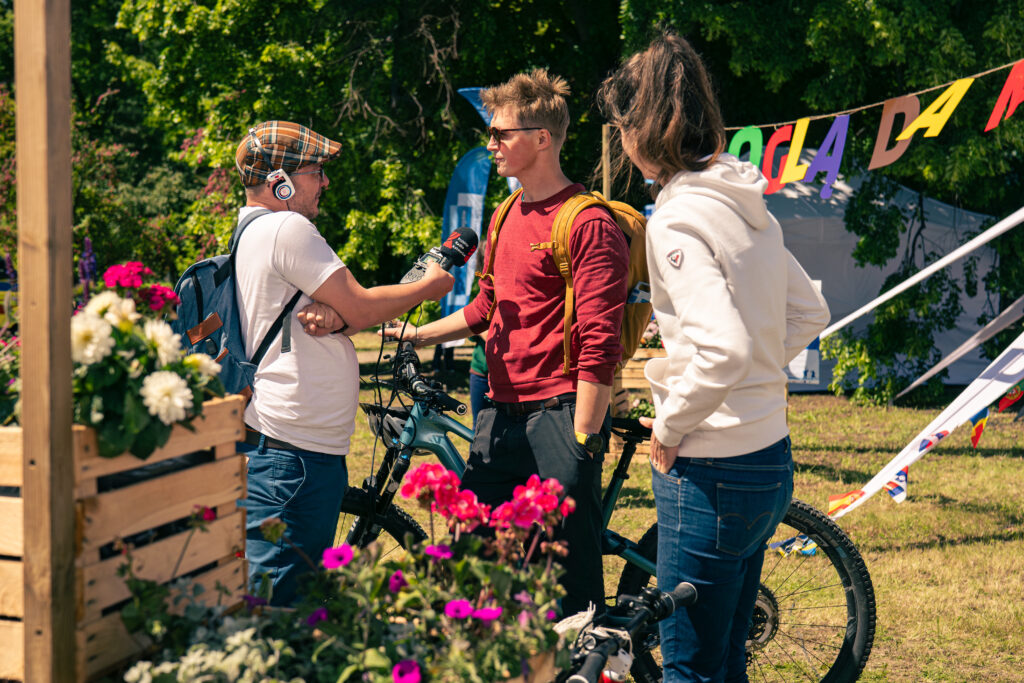 Reporter rozmawia z mężczyzną trzymającym rower. Obok stoi kobieta. Na pierwszym planie znajdują się skrzynki z kwiatami, a w tle namioty.