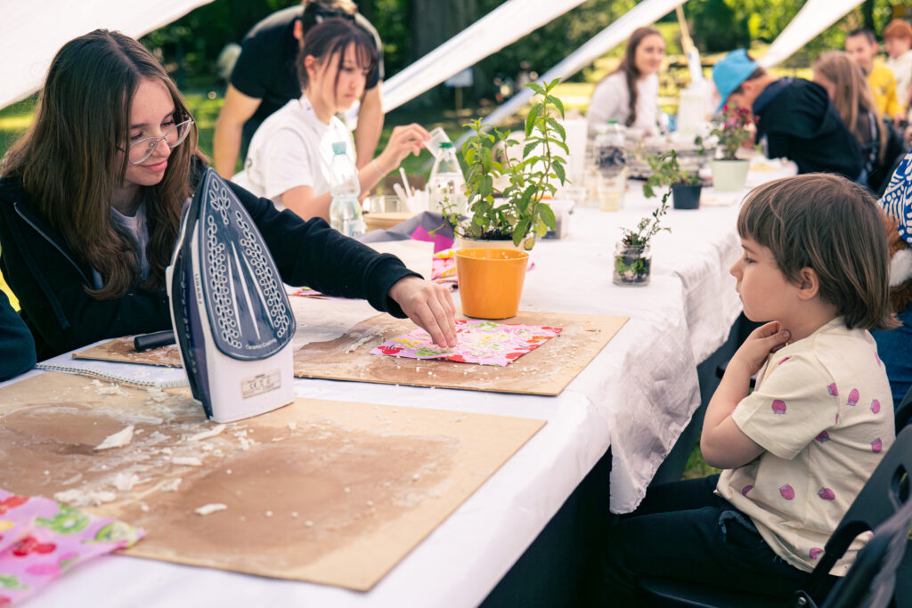 Zdjęcie przedstawia siedzących przy stołach animatorów oraz uczestników pikniku. Na pierwszym planie widoczne jest dziecko patrzące na pomagającą mu animatorkę. Na stole stoi żelazko i leży wiele kawałków wosku, które nakładane są na ozdobione wzorem kawałki materiału.