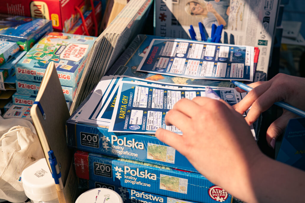 Na stosie puzzli leży karta gry, którą ktoś wypełnia. W tle widoczne są gry oraz gadżety.