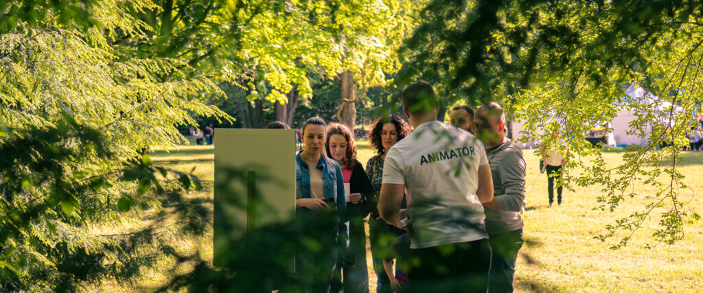 Na pierwszym planie widoczne są gałęzie drzew, za nimi znajduje się polana z namiotami. Przed animatorem stoi grupa ludzi.