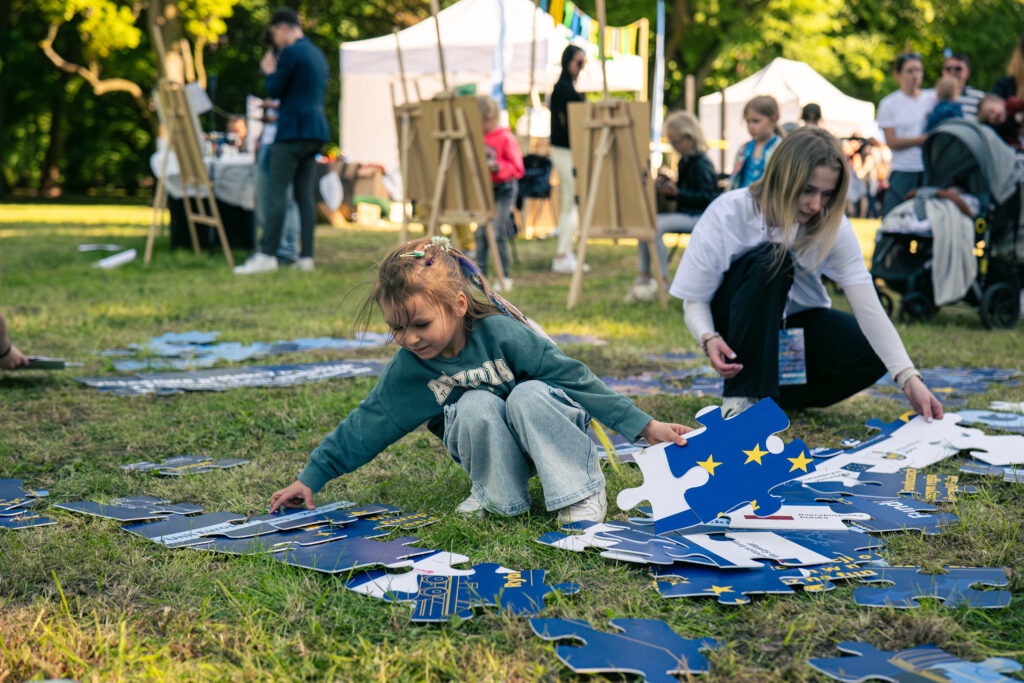 Uśmiechnięta dziewczynka układa na trawie ogromne puzzle, na których widoczne są fragmenty flagi Unii Europejskiej. Za nią znajduje się animatorka, a w tle widoczne są sztalugi, namioty oraz inni uczestnicy pikniku.