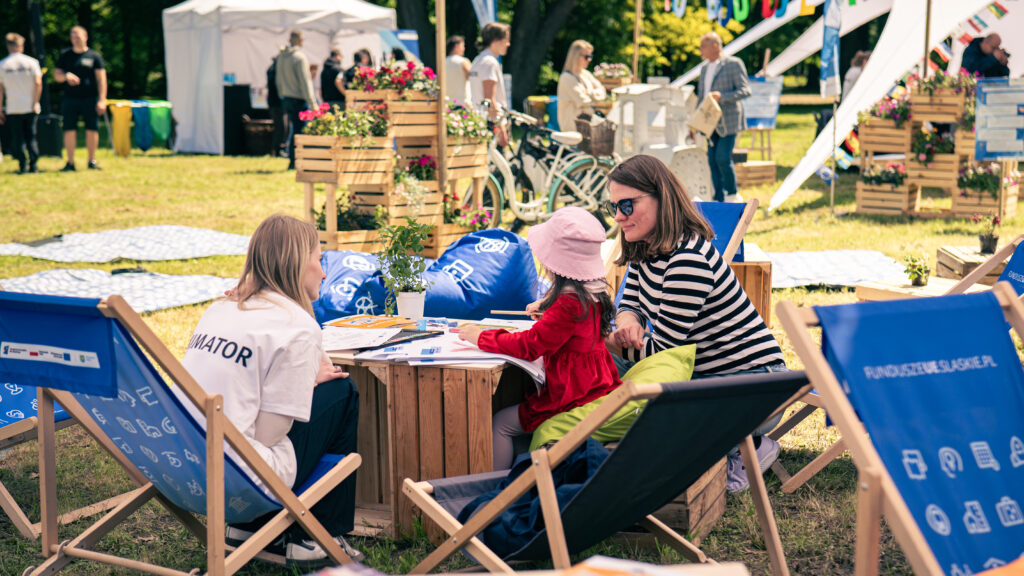 Na leżakach z napisami FunduszeUE.slaskie.pl siedzą kobiety. Jedna z nich jest animatorką. Obok stoi dziewczynka w kapeluszu oglądająca rozłożone na stole materiały reklamowe. W tle widoczni są inni uczestnicy pikniku, skrzynki z kwiatami oraz namioty.