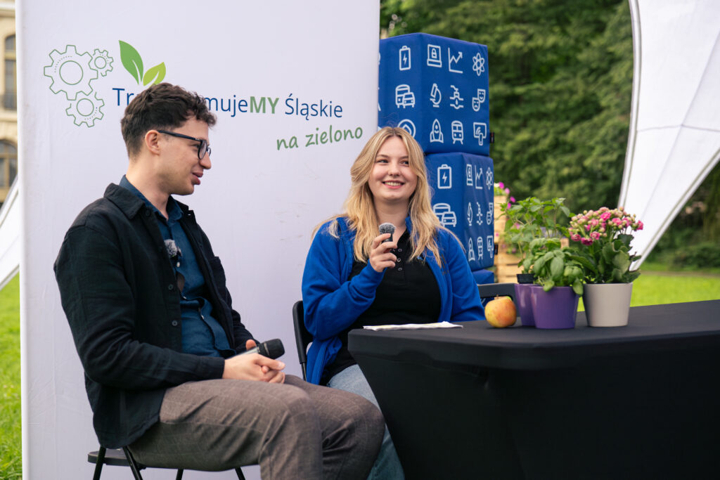 Uśmiechnięci kobieta i mężczyzna rozmawiają, siedząc przy stole, na którym znajdują się zioła i kwiaty w doniczkach oraz jabłko. W dłoniach trzymają mikrofony. Za nim znajduje się biała ścianka z napisem: TransformujeMY Śląskie na zielono.