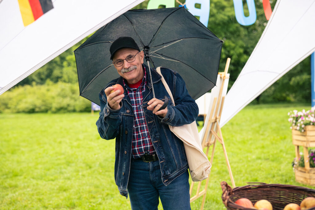 Uśmiechnięty mężczyzna w jednej dłoni trzyma rozłożony nad głową parasol, a w drugiej - jabłko.