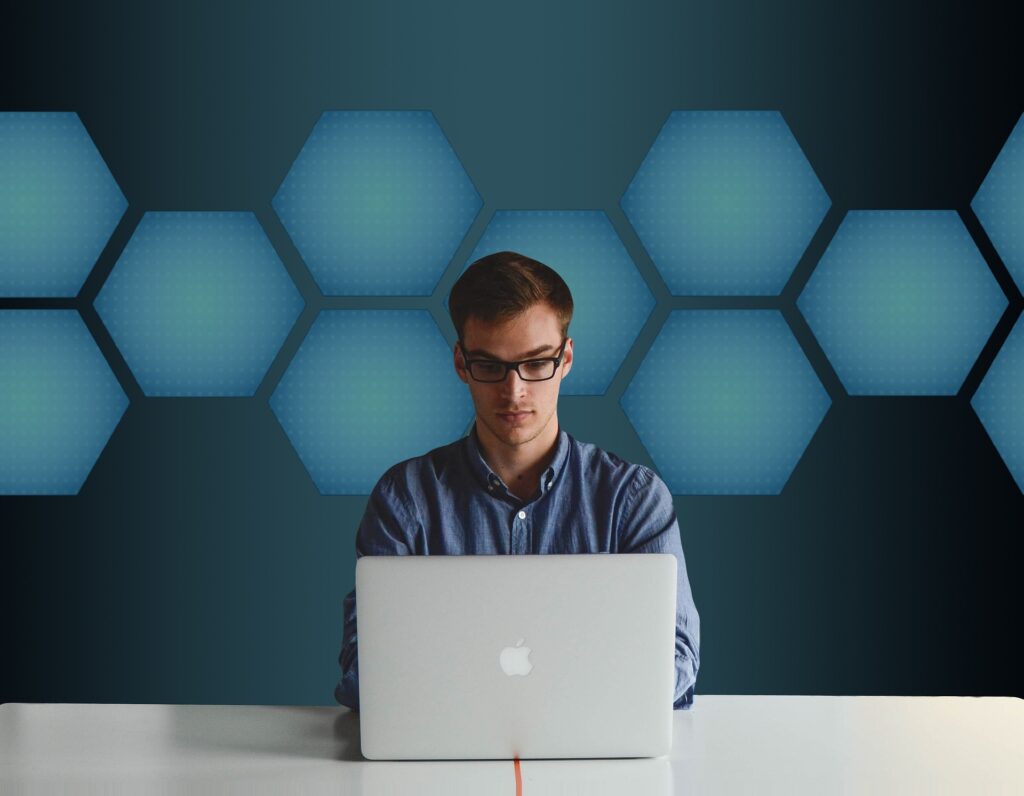 Zdjęcie przedstawia mężczyznę w okularach siedzącego przed długim białym blatem. Na blacie położony jest otwarty biały laptop. W tle widoczna jest ciemnoturkusowa ściana z pasem niebieskich sześciokątów.