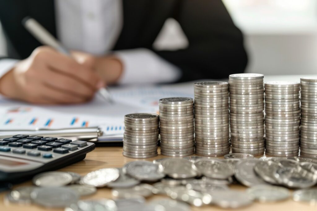 Na pierwszym planie biurka widoczne są stosy srebrnych monet. W tle znajduje się kalkulator i dokumenty. Siedząca za biurkiem osoba zaznacza coś w dokumentach.
