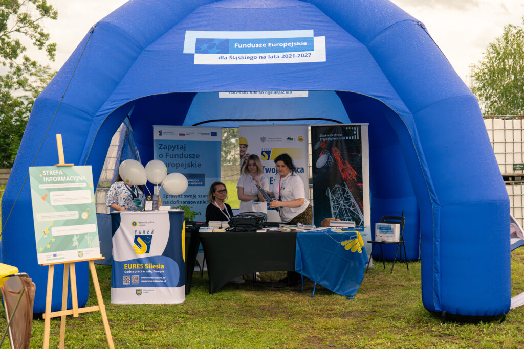 Zdjęcie przedstawia namiot z napisem Fundusze Europejskie dla Śląskiego na lata 2021-2027. W środku znajdują się stoiska, z których jedno ma napis Eures Silesia, oraz roll-upy z napisami: Śląskie Centrum Przedsiębiorczości, Zapytaj o Fundusze Europejskie. Stoiska obsługuje kilka kobiet.