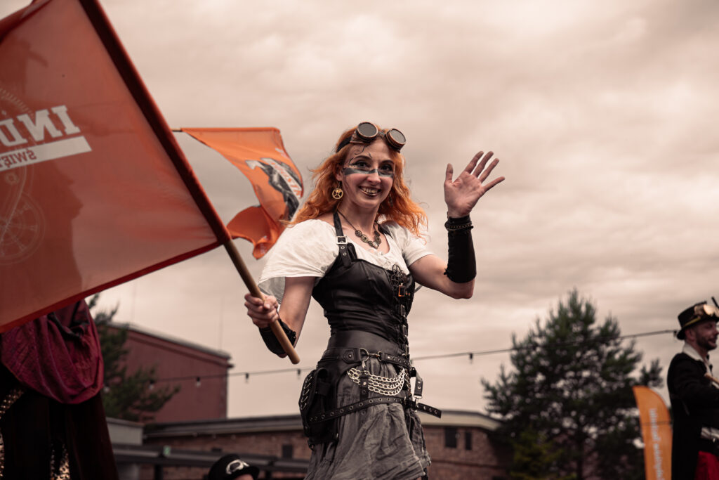 Zdjęcie przedstawia uśmiechniętą, machającą dłonią kobietę ubraną w stylu steampunk. W drugiej dłoni kobieta trzyma flagę, na której widoczny jest początek słowa Industriada.
