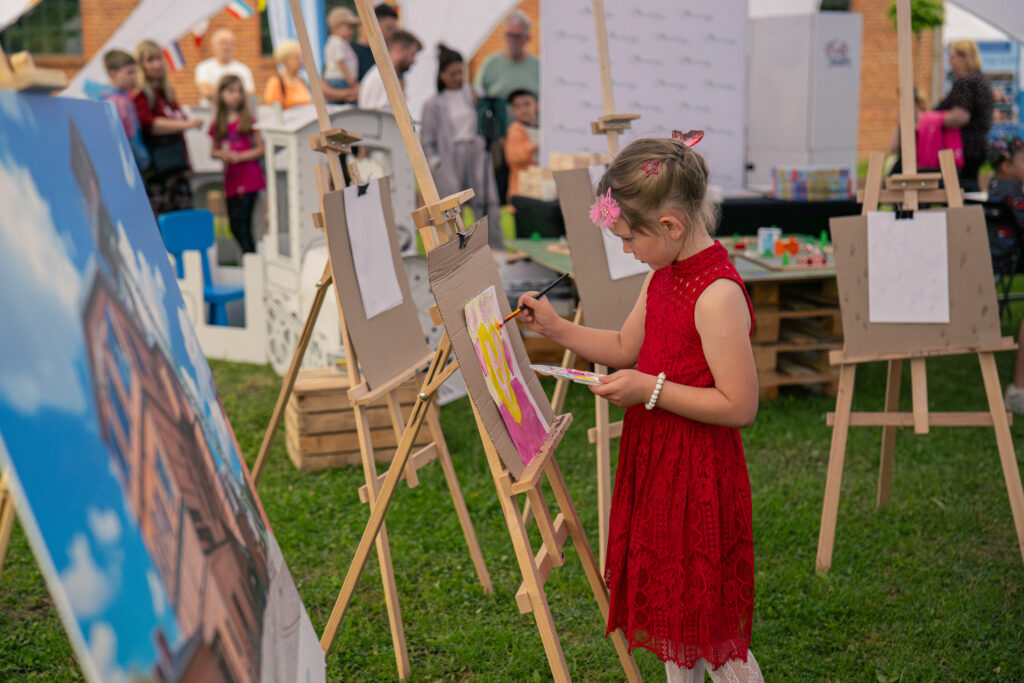 Dziewczynka stojąca przed sztalugami maluje obrazek. W tle widoczni są inni uczestnicy wydarzenia.