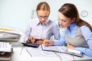 Zdjęcie przedstawia dwie kobiety siedzące przy biurku, na którym leży wiele dokumentów. Jedna z kobiet wskazuje ołówkiem z gumką dane na dokumencie, druga liczy na kalkulatorze.