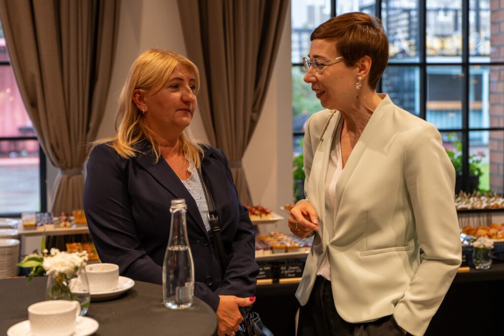 Zdjęcie ukazuje dwie uśmiechnięte kobiety rozmawiające przy stoliku. W tle widoczne są stoły z cateringiem oraz okna.