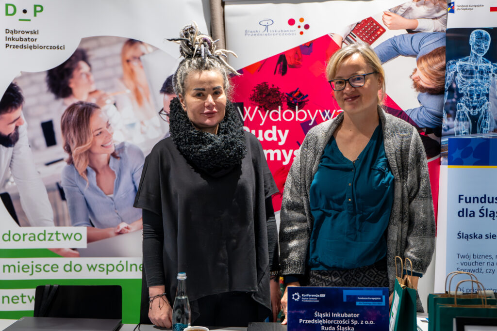 Dwie kobiety stoją obok siebie na stoisku informacyjnym. Kobieta po lewej ma fryzurę z dredami i ciemny, luźny strój. Kobieta po prawej, w okularach, jasnym kardiganie i turkusowej bluzce, uśmiecha się. Przed nimi leżą ulotki i butelka wody. W tle widoczne są plansze informacyjne promujące inkubatory przedsiębiorczości.