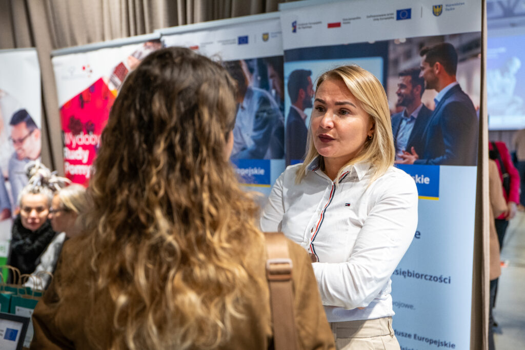 Bliskie ujęcie kobiety o blond włosach, ubranej w białą koszulę, rozmawiającej z drugą osobą, której widać tylko tył głowy i długie, falowane włosy. Kobieta w bieli patrzy na swoją rozmówczynię z uwagą. W tle znajdują się plansze informacyjne z tekstem i zdjęciami.