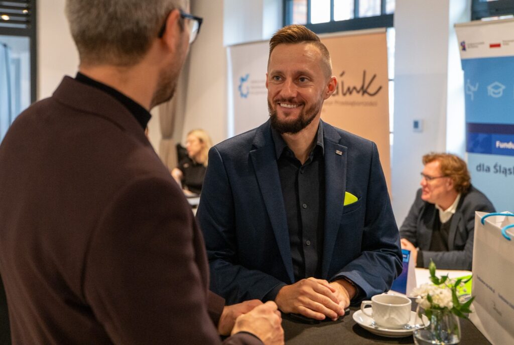 Zdjęcie przedstawia mężczyzn rozmawiających przy stoliku. Ten, który jest widoczny z przodu, szeroko się uśmiecha. W tle widoczne są stoiska informacyjne. Przy jednym z nich siedzi uśmiechnięty mężczyzna.