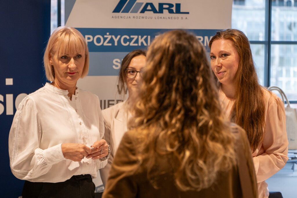 Zdjęcie przedstawia grupę uśmiechniętych kobiet rozmawiających przy stoisku informacyjnym. Na banerze widoczny jest napis: Agencja Rozwoju Lokalnego S.A.