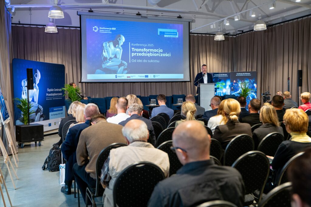 Szerokie ujęcie sali konferencyjnej. Mężczyzna stoi na podium, przemawiając do publiczności, która wypełnia rzędy krzeseł. W tle na ekranie wyświetla się tytuł "Konferencja 2025- Transformacja przedsiębiorczości - Od idei do sukcesu" oraz grafika przedstawiająca robota.