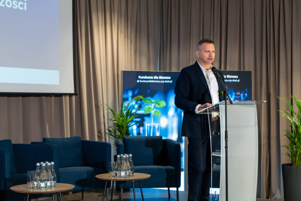 Mężczyzna w garniturze stoi za przezroczystą mównicą, przemawiając do publiczności. W tle widoczne są niebieskie fotele oraz plansza informacyjna promująca "Fundusze dla Biznesu" po prawej stronie.