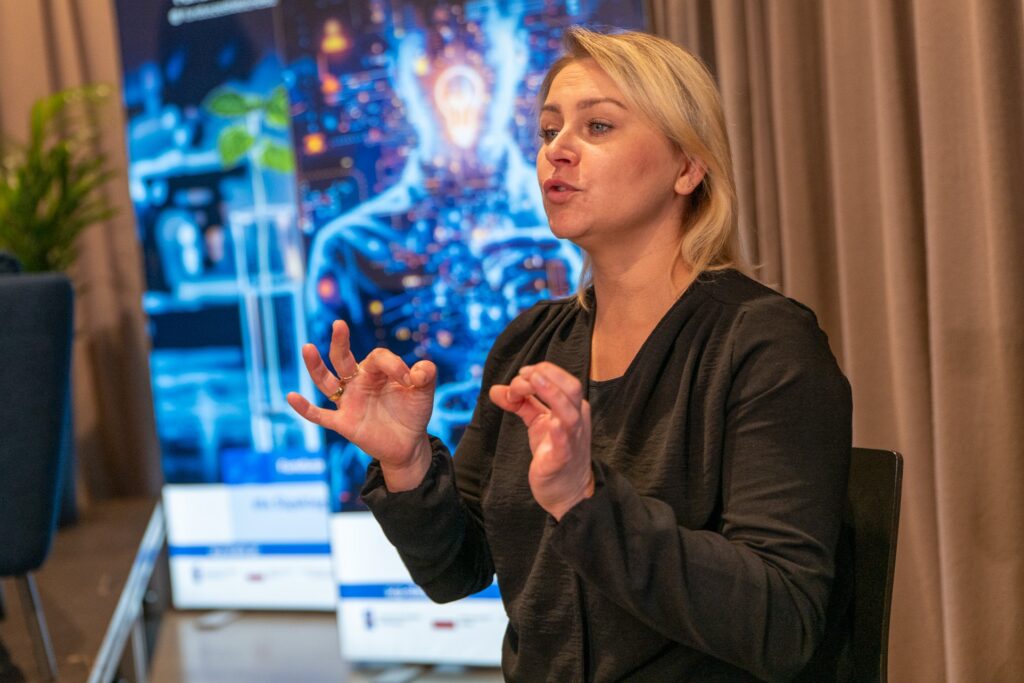 Zdjęcie przedstawia kobietę przemawiającą w języku migowym. W tle widoczne są podświetlne ścianki. Jedna z rośliną w próbówce, druga z motywami cyfrowymi i zarysem ludzkiej sylwetki.