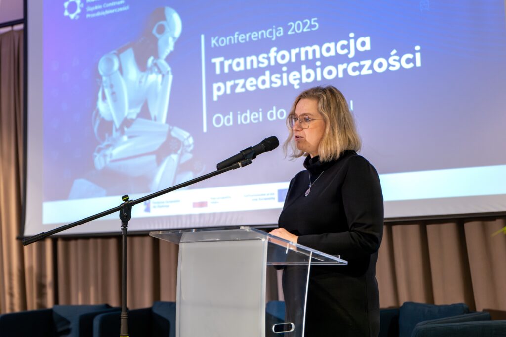 Kobieta w okularach, ubrana w ciemny golf, stoi za mównicą, przemawiając do mikrofonu. Na dużym ekranie za nią widnieje napis "Konferencja 2025 Transformacja przedsiębiorczości" i grafika przedstawiająca robota.