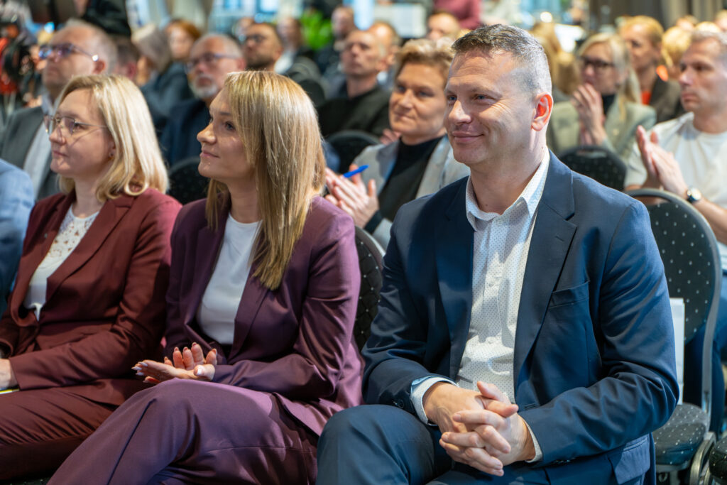 Trzy osoby (dwie kobiety i mężczyzna) siedzą na widowni konferencji. Wszyscy są elegancko ubrani i patrzą w lewo, skupieni na scenie. W tle widoczna jest klaszcząca publiczność.