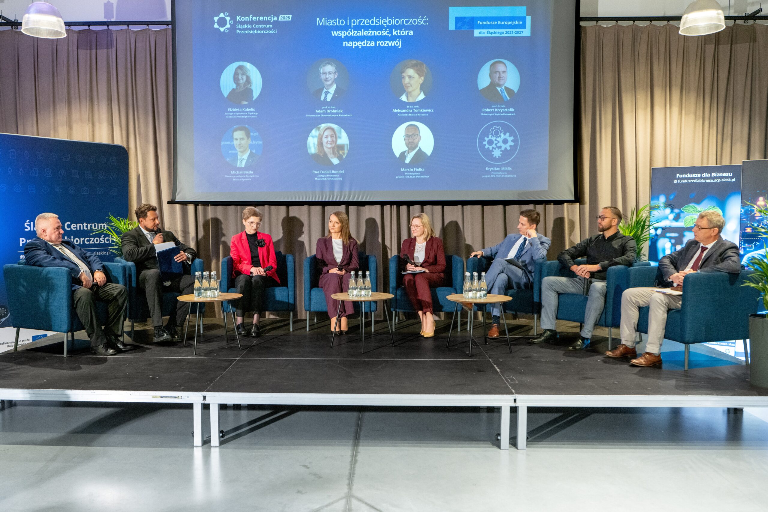 Zdjęcie przedstawia panel dyskusyjny podczas konferencji. Dziewięć osób siedzi na niebieskich fotelach na podwyższeniu sceny. W tle duży ekran wyświetla prezentację z tytułem 