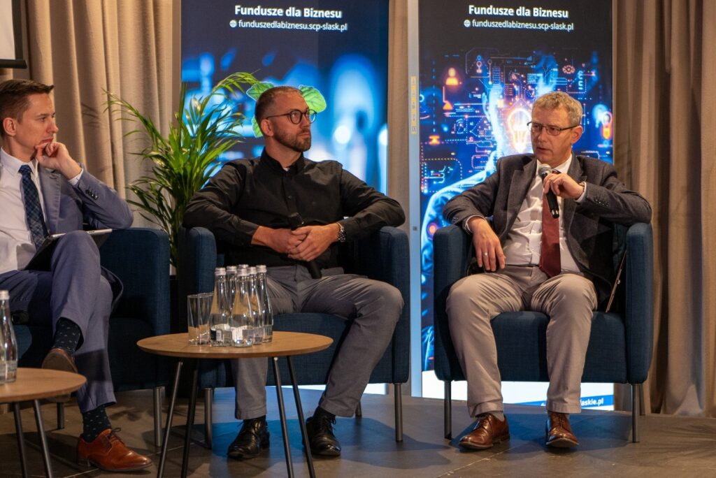 Zdjęcie ukazuje trzech mężczyzn siedzących w fotelach. Jeden z nich trzyma mikrofon. W tle widoczne są podświetlone ścianki z reklamą strony funduszedlabiznesu.scp-slask.pl oraz rośliny. Na pierwszym planie znajduje się stolik ze stojącymi na nim butelkami wody.