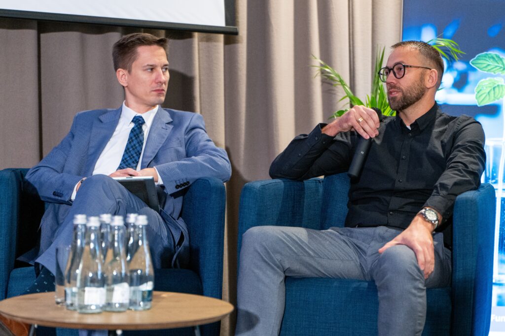 Dwóch mężczyzn siedzi na niebieskich fotelach na scenie. Mężczyzna po prawej, w czarnej koszuli i okularach, rozmawia, trzymając mikrofon. Mężczyzna po lewej, w garniturze, słucha. Przed nimi na stoliku stoją butelki wody.