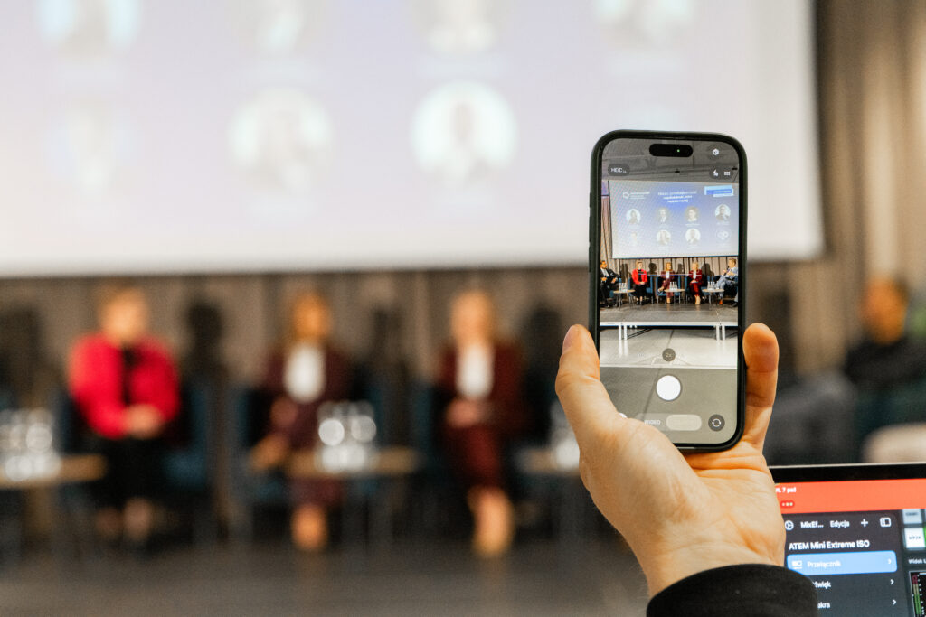 Zbliżenie na dłoń trzymającą smartfon. Ekran telefonu wyświetla podgląd nagrywanego panelu dyskusyjnego na scenie. Tło jest rozmyte, co skupia uwagę na urządzeniu i rejestrowanym obrazie.