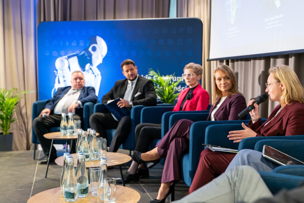 Fragment panelu dyskusyjnego. Pięć osób (w tym trzy kobiety i dwóch mężczyzn) siedzi na niebieskich fotelach. Kobieta w bordowym garniturze i okularach przemawia do mikrofonu.