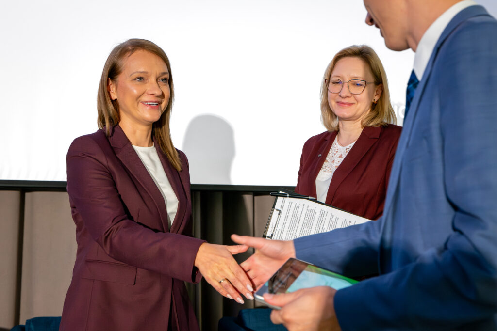 Trzy osoby podczas uścisku dłoni na scenie. Kobieta w bordowym garniturze po lewej uśmiecha się, witając się z mężczyzną widocznym częściowo po prawej. Druga kobieta w okularach, również w bordowym garniturze, stoi z tyłu i przygląda się z uśmiechem. W tle widać biały ekran.