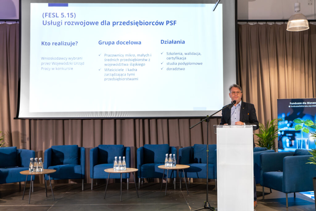 Mężczyzna w okularach i marynarce stoi za mównicą i przemawia podczas konferencji. W tle na dużym ekranie wyświetlany jest slajd informujący o usługach rozwojowych dla przedsiębiorców PSF, z podziałem na sekcje: "Kto realizuje?", "Grupa docelowa" i "Działania" w języku polskim. W głębi sceny znajdują się niebieskie fotele i małe stoliki z butelkami wody.