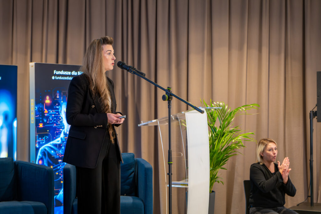 Kobieta o długich włosach, ubrana w czarny garnitur, stoi obok przezroczystej mównicy i przemawia, trzymając w dłoni małego pilota. Po prawej stronie, niżej, siedzi druga kobieta, wykonująca tłumaczenie na język migowy. W tle widoczna jest duża plansza informacyjna i brązowa zasłona.