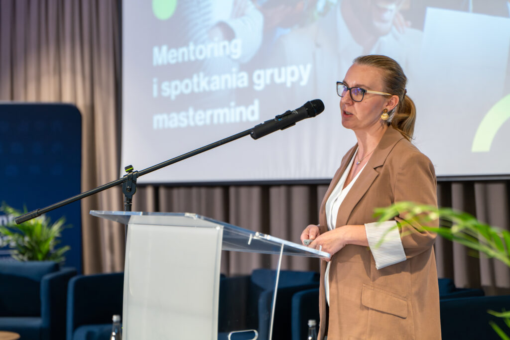 Kobieta w okularach i beżowej marynarce przemawia do mikrofonu, stojąc za przezroczystą mównicą. W tle na ekranie wyświetla się tekst "Mentoring i spotkania grupy mastermind".