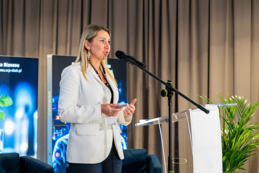 Kobieta o jasnych włosach, ubrana w białą marynarkę, przemawia do mikrofonu za przezroczystą mównicą. W tle widoczne są duże plansze informacyjne i rośliny doniczkowe.