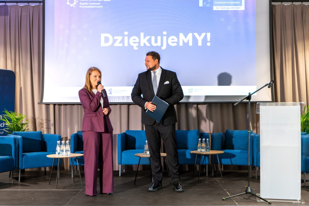 Dwie osoby stoją na scenie przed dużym ekranem z napisem "Dziękujemy!". Kobieta po lewej, ubrana w bordowy garnitur, trzyma mikrofon i przemawia. Mężczyzna po prawej, w ciemnym garniturze i z teczką w ręku, stoi obok. W tle widoczne są niebieskie fotele i małe stoliki z wodą.