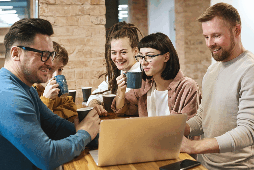 Grupa pięciu osób siedzi wokół drewnianego stołu w pomieszczeniu o ciepłym oświetleniu. Piją kawę lub herbatę z kubków i z uśmiechami patrzą na ekran laptopa leżącego na stole.