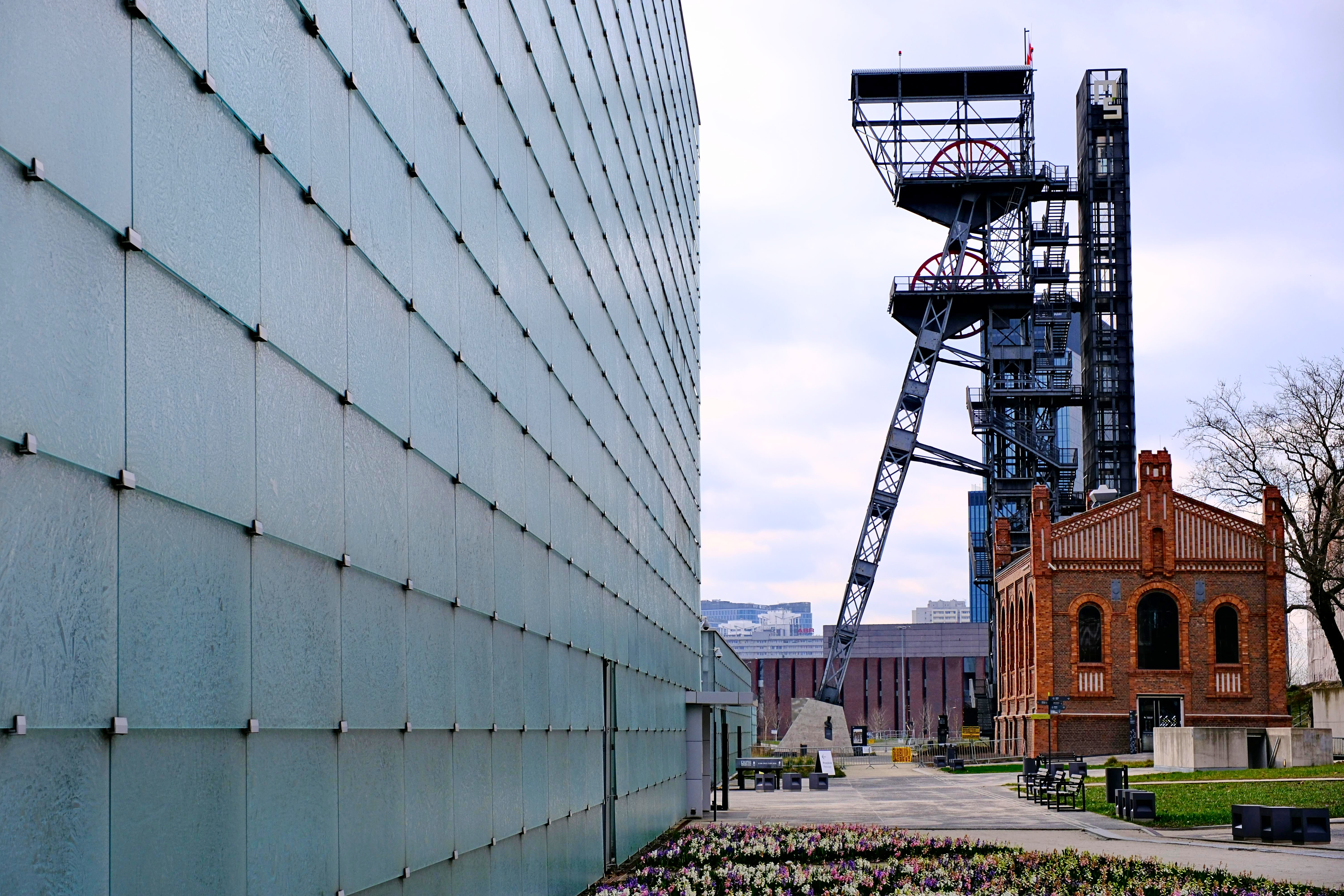 Połączenie nowoczesnej i historycznej architektury. Po lewej stronie dominuje duża, przeszklona fasada Muzeum Śląskiego, a po prawej widać czerwoną, ceglaną zabudowę i zabytkową stalową wieżę wyciągową szybu kopalnianego (Szyb Warszawa II) w Katowicach.
