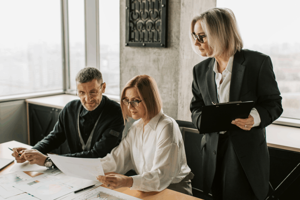 Kobieta i mężczyzna siedzą przy stole w nowoczesnym biurze, przeglądając dokumenty i wykresy. kobieta w garniturze stoi obok, trzymając podkładkę pod notatki i spoglądając na materiały.