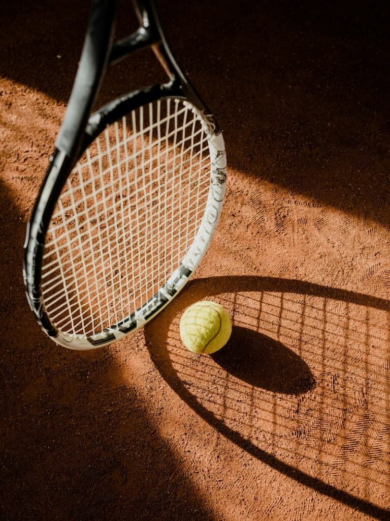 Rakieta i piłka tenisowa na korcie ziemnym, rzucające geometryczny cień, co symbolizuje infrastrukturę sportową. Takie obiekty, powstające na terenach poprzemysłowych, mogą pomóc inwestorowi zdobyć punkty za zagospodarowanie terenu o powierzchni powyżej 1 hektara.