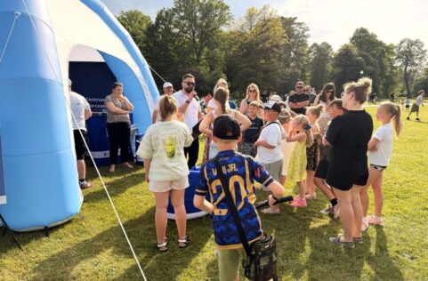 Zdjęcie z Pikniku Europejskiego przedstawia grupę dzieci i dorosłych zgromadzonych na słonecznej polanie obok dużego, niebiesko-białego namiotu sferycznego. Pośrodku stoi prowadzący w białej koszulce i okularach przeciwsłonecznych, który mówi do mikrofonu, animując zebranych uczestników. Na pierwszym planie, tyłem do obiektywu, stoi chłopiec w sportowej, niebiesko-złotej koszulce piłkarskiej z numerem 20 i z przewieszoną przez ramię torbą. W tle widać liczną publiczność stojącą na zielonej trawie oraz ścianę wysokich drzew w pełnym słońcu.